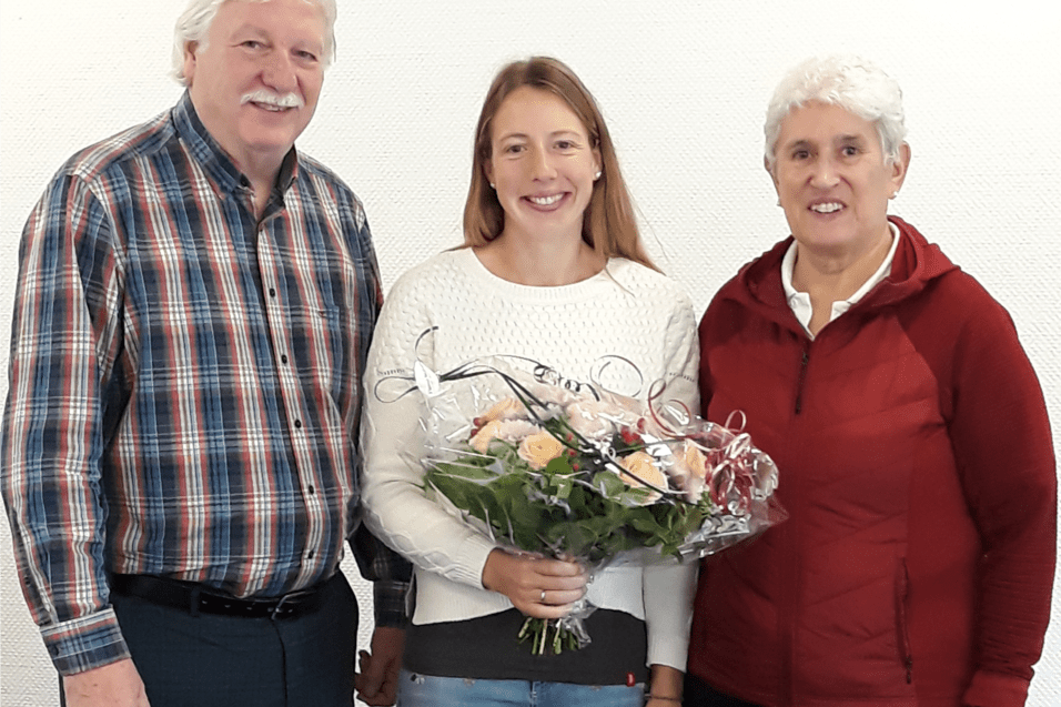 Drei Personen posieren vor einem wei&szlig;en Hintergrund, eine Frau h&auml;lt einen Blumenstrau&szlig;.