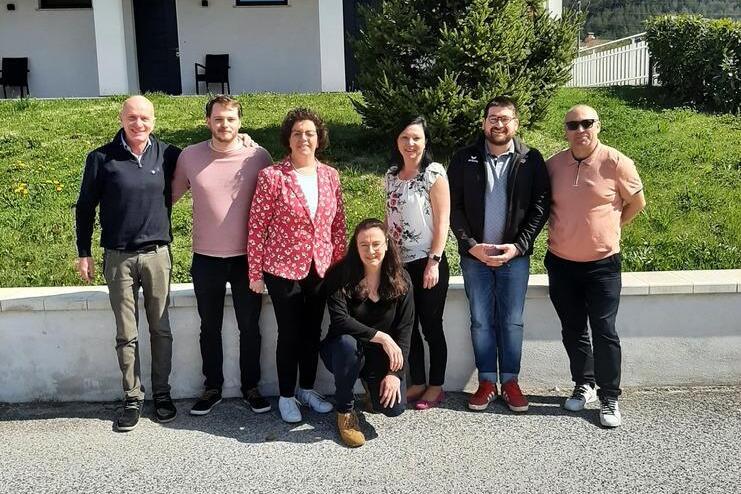 Gruppe von sechs Personen, die vor einem Haus mit einem Baum stehen, blauer Himmel und grüne Wiese im Hintergrund.