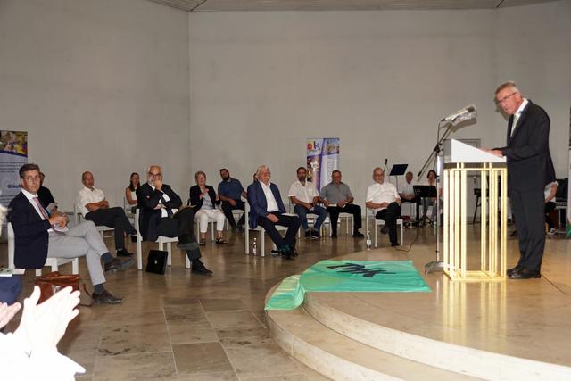 Mann am Rednerpult vor Publikum in einem hellen Raum, das an einer Veranstaltung teilnimmt.