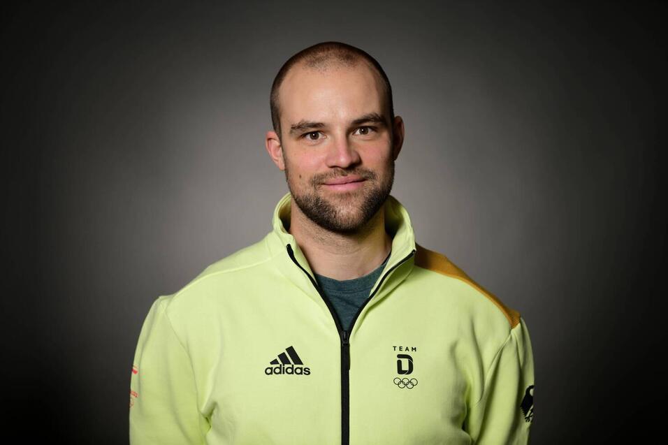 Portr&auml;t eines Mannes mit kurzen Haaren, der eine hellgr&uuml;ne Jacke mit olympischem Logo tr&auml;gt, vor einem dunklen Hintergrund.