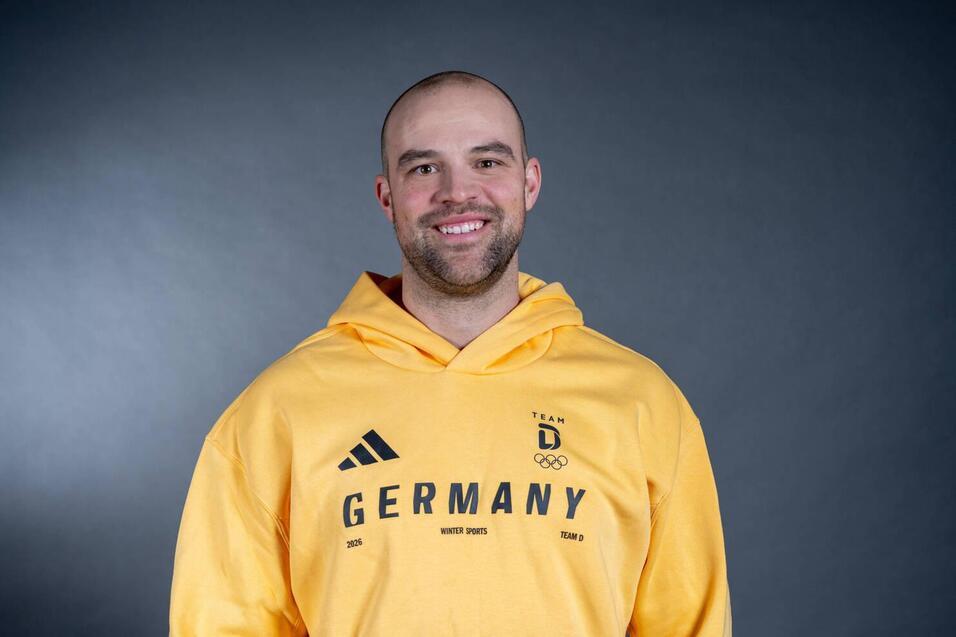 L&auml;chelnder Mann in gelbem Olympiatrainingsanzug mit Aufschrift &bdquo;Germany&ldquo; vor grauem Hintergrund.