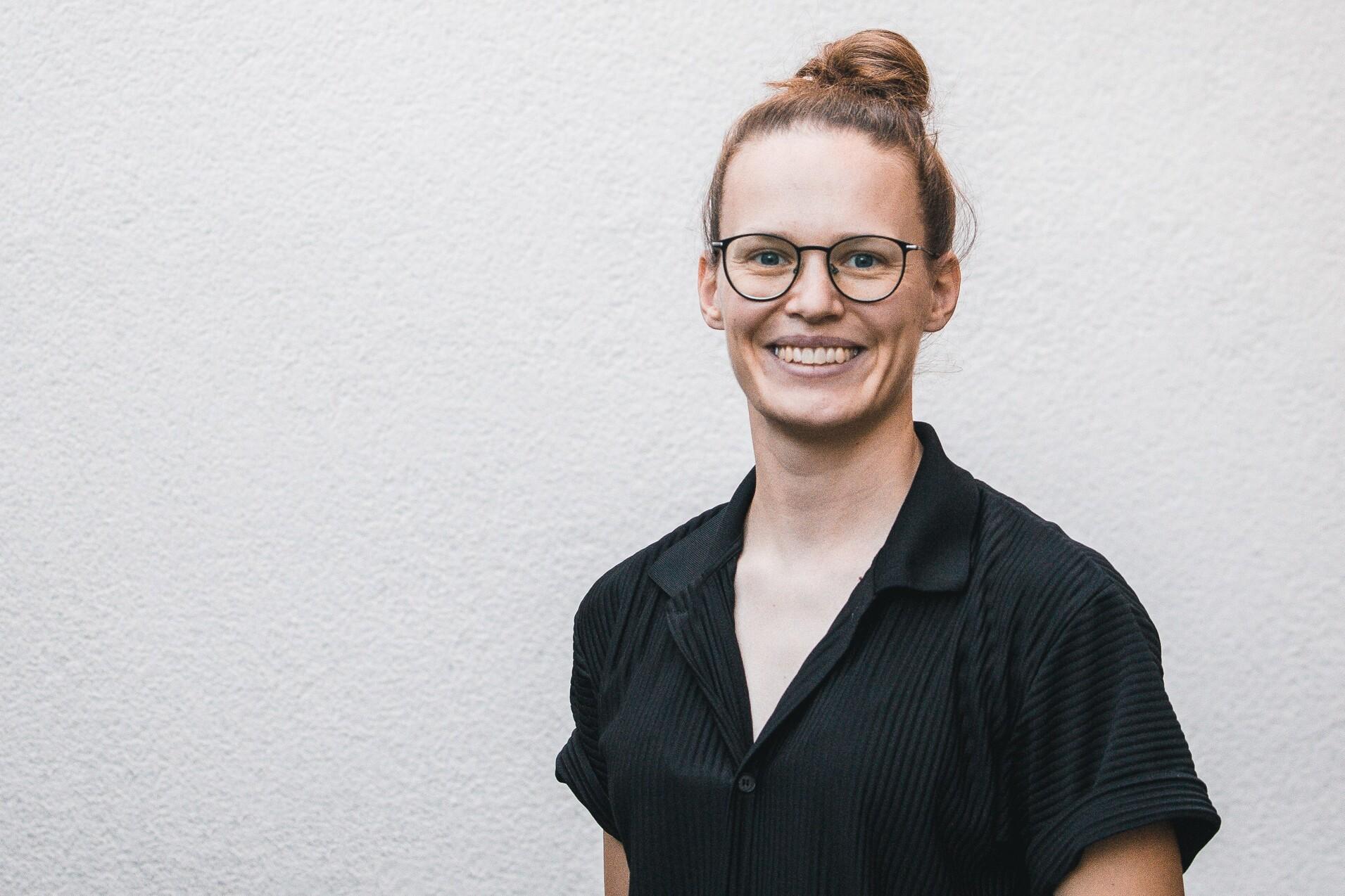 Frau mit schulterlangen, braunen Haaren im Dutt, tr&auml;gt eine schwarze Bluse und Brille, steht vor einer hellen Wand.