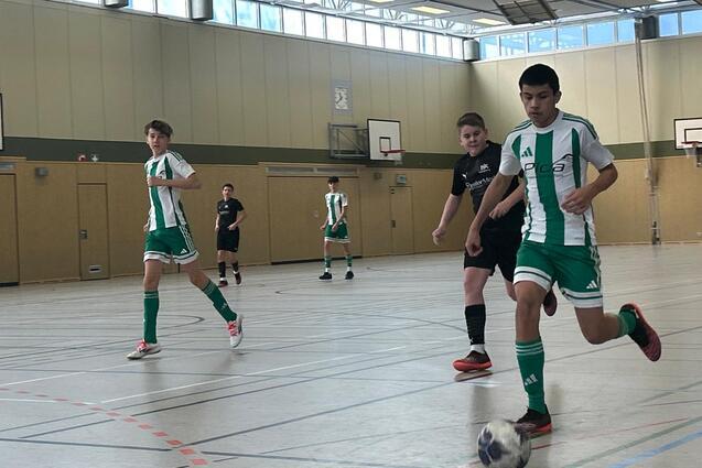 Drei Jugendliche spielen Fu&szlig;ball in einer Sporthalle, einer f&uuml;hrt den Ball, w&auml;hrend die anderen verfolgen.
