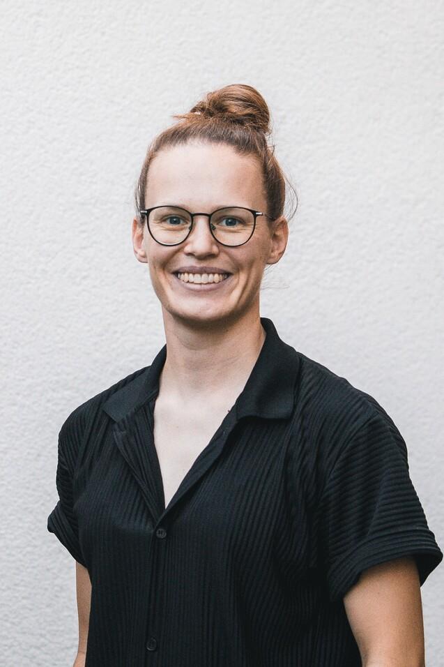 Frau mit schulterlangen, braunen Haaren im Dutt, tr&auml;gt eine schwarze Bluse und Brille, steht vor einer hellen Wand.