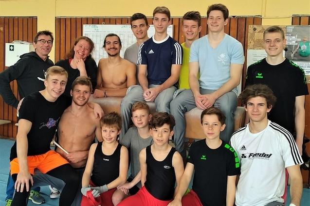 Gruppe von Jugendlichen und Trainern l&auml;chelt auf einem Gymnastikboden, umgeben von Sportger&auml;ten und einer Wandtafel.