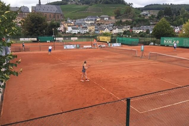 Tennisplatz mit Spielern auf roter Asche, umgeben von typischer Architektur und Weinbergen im Hintergrund.