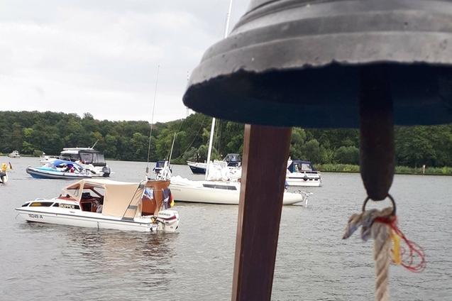 Nahaufnahme einer Schiffsglocke im Vordergrund, mit Booten und Yachten auf einem Gew&auml;sser im Hintergrund.