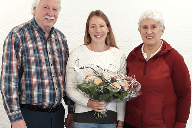 Drei Personen posieren vor einem wei&szlig;en Hintergrund, eine Frau h&auml;lt einen Blumenstrau&szlig;.