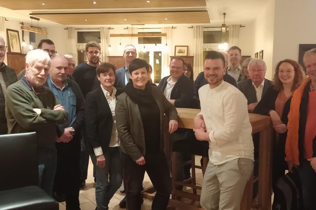 Gruppe von 16 Personen in einem Restaurant, l&auml;chelnd vor einer Bartheke, mit dekorativen Bildern im Hintergrund.