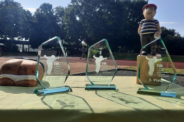 Drei gl&auml;serne Pokale auf einem Tisch, mit einem Holzfigur und einem Leckerli im Hintergrund, unter blauem Himmel.