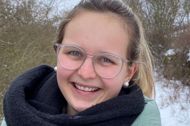 L&auml;chelnde junge Frau mit Brille und Ohrringen, eingepackt in einen schwarzen Schal, beim Spaziergang im Schnee.