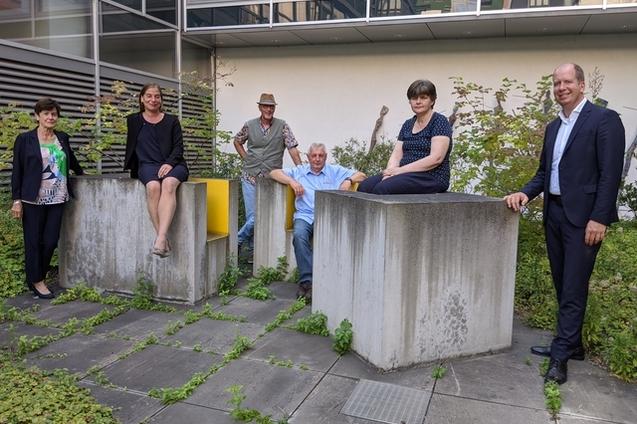 Sechs Personen posieren auf modernen Betonblock-Sitzen in einem begr&uuml;nten Innenhof eines Geb&auml;udes.