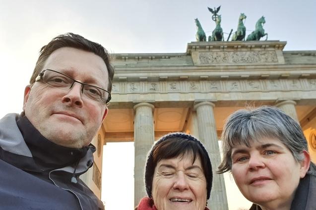 Drei Personen posieren vor dem Brandenburger Tor im Hintergrund, bei Sonnenaufgang in Berlin.