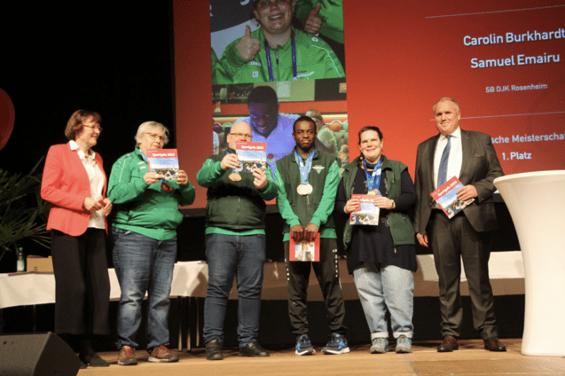 Gruppe von Preistr&auml;gern mit Medaillen und Urkunden auf der B&uuml;hne bei einer Veranstaltung, mit Rednern im Hintergrund.