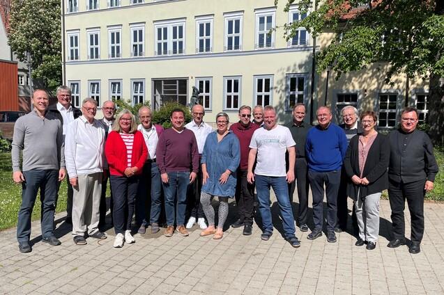 Gruppe von 18 Personen steht auf einem gepflasterten Platz vor einem mehrst&ouml;ckigen Geb&auml;ude mit Fenstern.