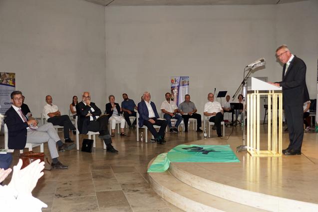 Mann am Rednerpult vor Publikum in einem hellen Raum, das an einer Veranstaltung teilnimmt.