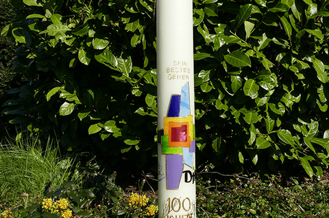 Bunte S&auml;ule mit dem Schriftzug &bdquo;100 Jahre 1920&ldquo; und grafischen Elementen, vor einer gr&uuml;nen Hecke im Freien.