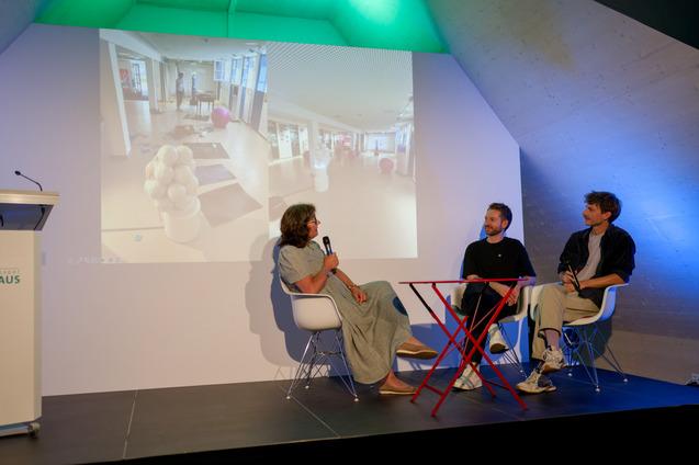 Drei Personen sitzen an einem Tisch und diskutieren, w&auml;hrend eine Pr&auml;sentation im Hintergrund gezeigt wird.