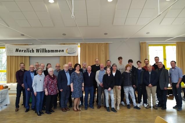 Gruppenfoto von 27 Personen in einem Raum, in dem ein Willkommensbanner h&auml;ngt. Alle l&auml;cheln in die Kamera.