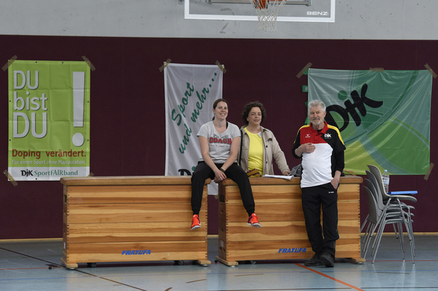 Drei Personen sitzen und stehen neben einer Holzwand im Sportraum, umgeben von Werbebannern und St&uuml;hlen.