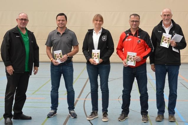 F&uuml;nf Personen stehen in einer Sporthalle, halten Brosch&uuml;ren und l&auml;cheln in die Kamera.