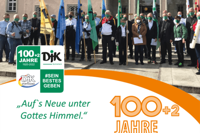 Gruppenshot von Personen in Masken, die Fahnen halten, vor einem Geb&auml;ude mit Jubil&auml;umsbanner &bdquo;100+2 Jahre&ldquo;.