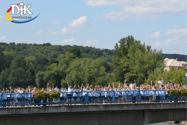 Gruppenszene auf einer Br&uuml;cke mit vielen Menschen, die H&auml;nde heben, umgeben von gr&uuml;nen B&auml;umen und blauen Himmel.