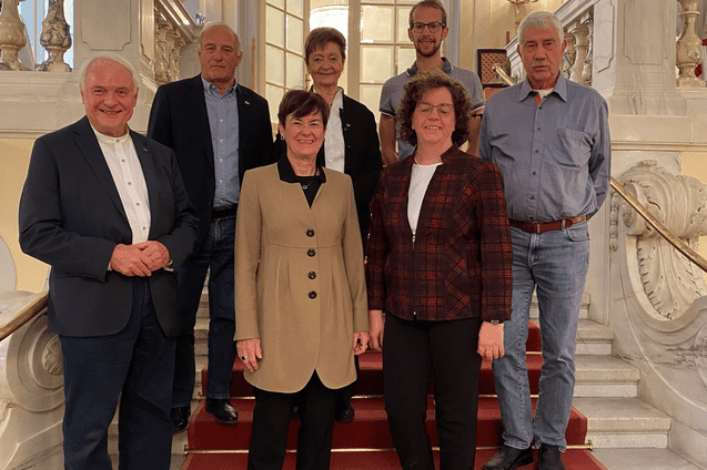 Gruppe von sieben Personen auf einer Treppe mit roten Teppichen in einem stilvollen Geb&auml;ude.