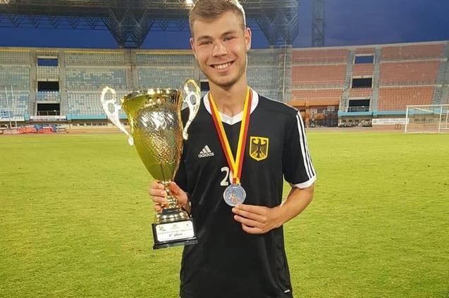 Ein junger Fu&szlig;ballspieler in Schwarz h&auml;lt einen Pokal und eine Medaille auf einem gr&uuml;nen Rasenplatz in einem Stadion.