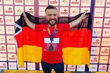 L&auml;chelnder Athlet in rot-schwarzem Trikot h&auml;lt die deutsche Flagge und eine Medaille vor einer Sponsorenwand.