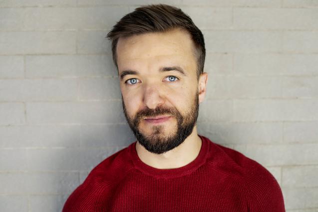 Portr&auml;t eines Mannes mit Kurzhaarfrisur und Bart, l&auml;chelnd vor einer grauen Backsteinwand.