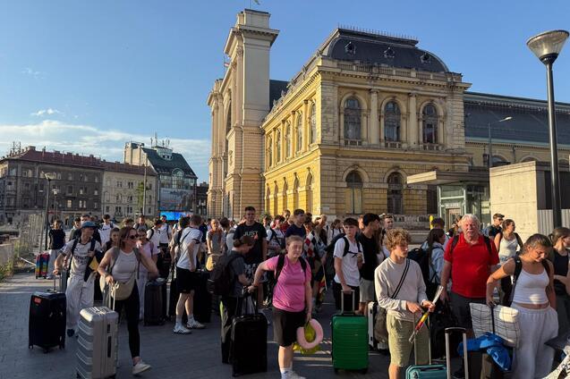 Reihe von Reisenden mit Gep&auml;ck vor einem historischen Bahnhof bei Sonnenlicht.