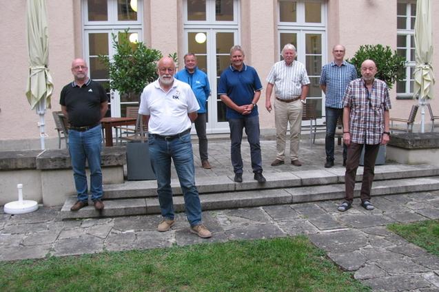 Gruppe von sieben M&auml;nnern steht auf einerTerrasse vor einem Geb&auml;ude, umgeben von B&auml;umen und St&uuml;hlen.