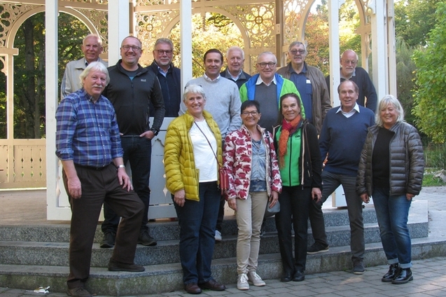 Gruppe von 15 Personen steht vor einem Pavillon in einem Park, umgeben von B&auml;umen und Gr&uuml;nfl&auml;chen.