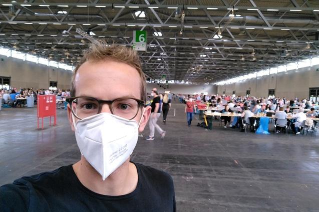 Selbstportr&auml;t eines Mannes mit Maske in einer gro&szlig;en Halle mit vielen Tischen und Personen bei einer Veranstaltung.
