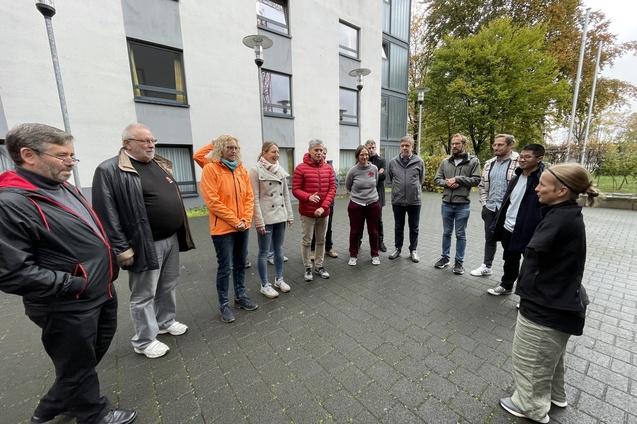 Gruppe von zehn Personen steht im Freien, umgeben von B&auml;umen und Geb&auml;uden, w&auml;hrend eine Person spricht.