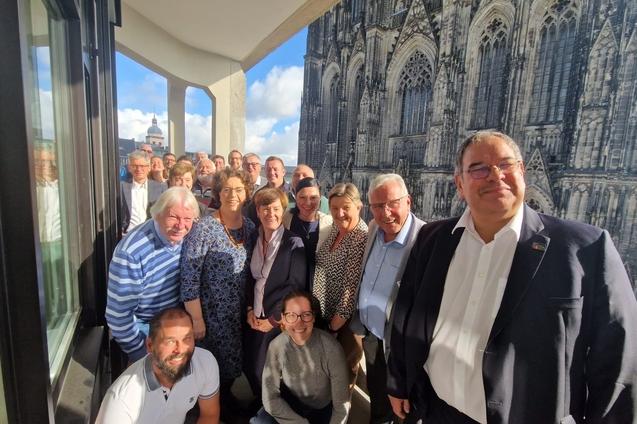 Gruppe von 20 Personen posiert l&auml;chelnd vor einer gro&szlig;en Fensterfront mit Blick auf eine Kathedrale.