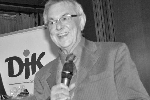 L&auml;chelnder Mann mit Mikrofon steht vor einem DJK-Logo und spricht an einem Podium.