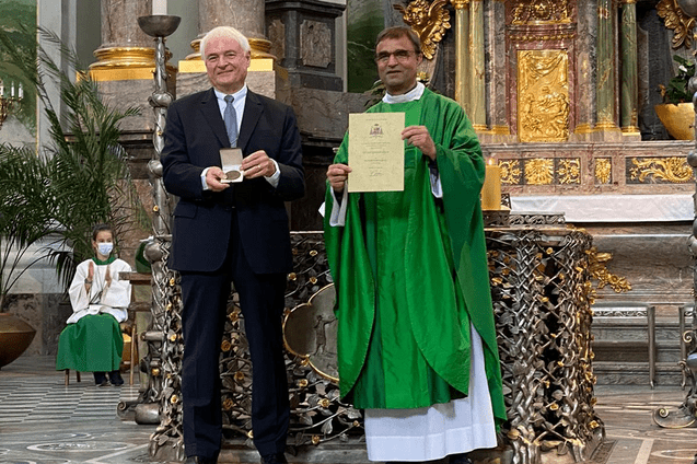 Zwei M&auml;nner stehen in einer Kirche; einer h&auml;lt eine Urkunde, der andere eine Medaille, im Hintergrund eine Person in Kirchenkleidung.