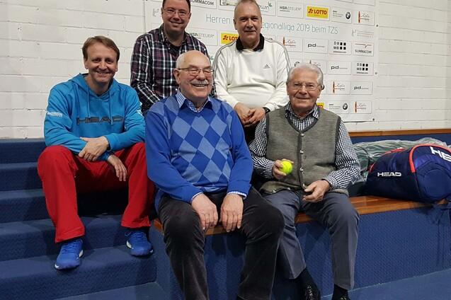 F&uuml;nf M&auml;nner sitzen und stehen auf einer Holztrib&uuml;ne, drei halten einen Tennisball, im Hintergrund Werbefl&auml;chen.