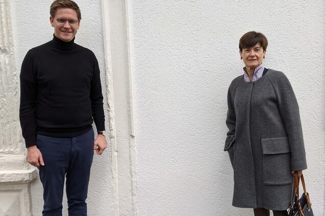 Zwei Personen stehen vor einer wei&szlig;en Wand: ein Mann in schwarzem Pullover und eine Frau in grauem Mantel.