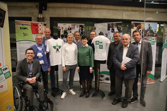 Gruppenfoto von zehn Personen, die vor einer Ausstellung mit Sportdress und Informationsplakaten stehen.