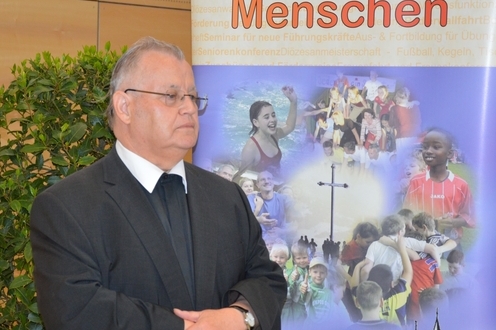 Ein Mann in Anzug steht neben einem Plakat mit dem Slogan &bdquo;Sport bewegt Menschen&ldquo; und Bildern sportlicher Aktivit&auml;ten.