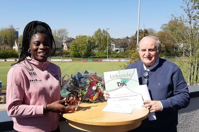 Eine junge Sportlerin h&auml;lt einen Preis und eine Urkunde, w&auml;hrend ein &auml;lterer Mann daneben steht, auf einem Sportplatz.