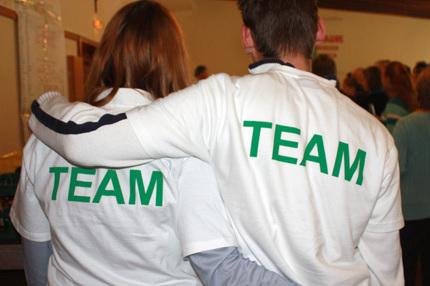 Zwei Personen mit R&uuml;ckenansicht tragen wei&szlig;e T-Shirts mit gr&uuml;ner Aufschrift &bdquo;TEAM&ldquo; auf ihren R&uuml;cken.