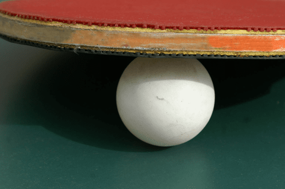 Tischtennisschl&auml;ger mit roter Oberfl&auml;che schwebt &uuml;ber einem wei&szlig;en Tischtennisball auf gr&uuml;nem Tisch.