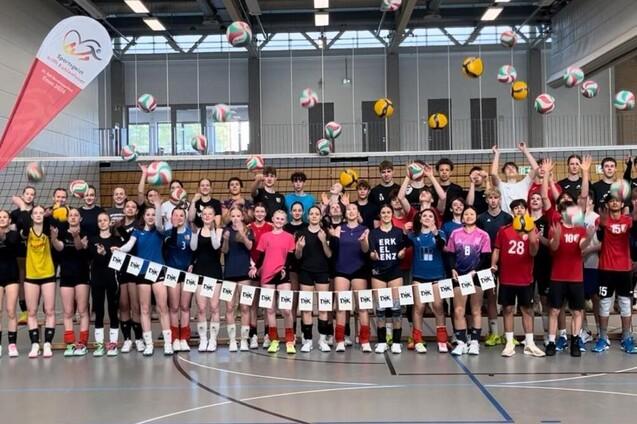 Gruppenszene von Volleyballspielern, die mit B&auml;llen in der Luft jubeln, vor einem Volleyballnetz in einer Halle.