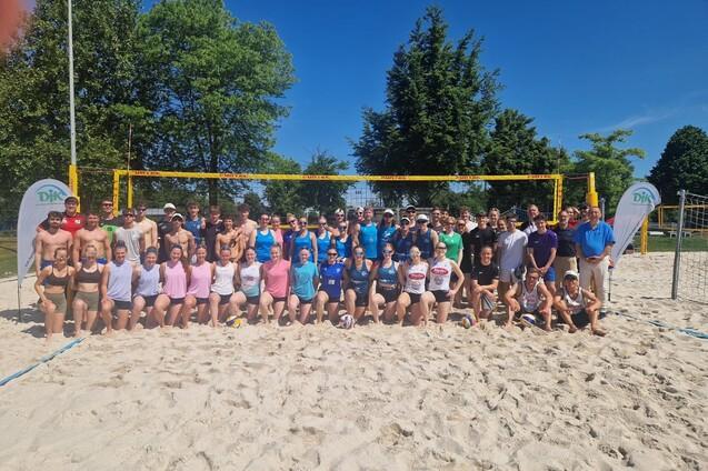 Gruppenshot von 50 Beachvolleyballspielern auf einem Sandplatz, umrahmt von B&auml;umen und einem blauen Himmel.