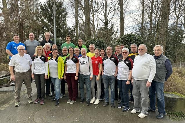 Gruppenfoto von 25 Personen in Sportkleidung, stehend in einem Park mit B&auml;umen und Laternen im Hintergrund.