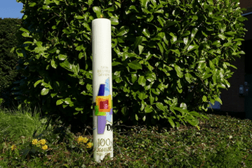 S&auml;ule mit einem bunten Design und dem Aufdruck "100 Jahre" vor einer gr&uuml;nen Hecke in sonniger Umgebung.